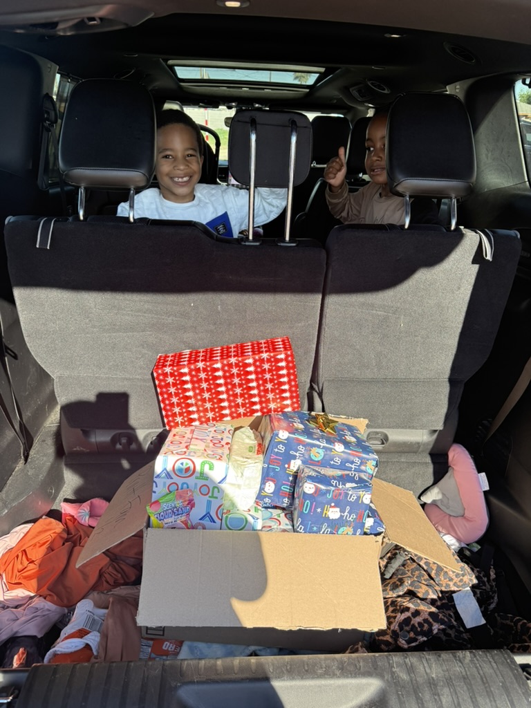Two boys smiling from a car with a box of wrapped presents visible in the trunk