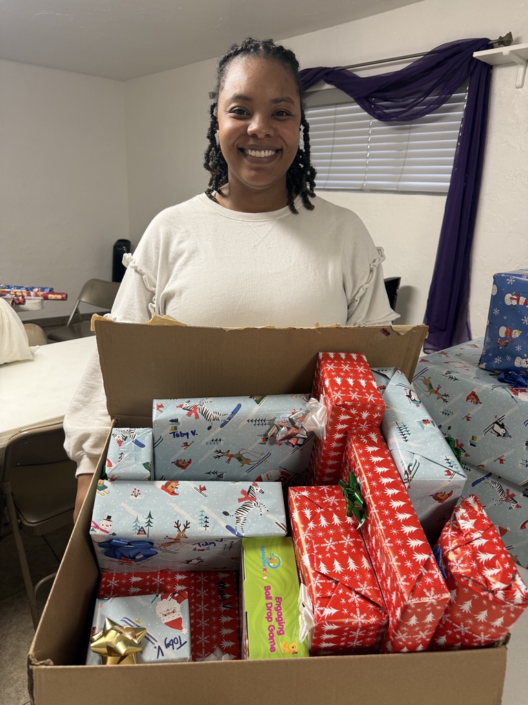 A smiling woman in white holding a box filled with wrapped gifts