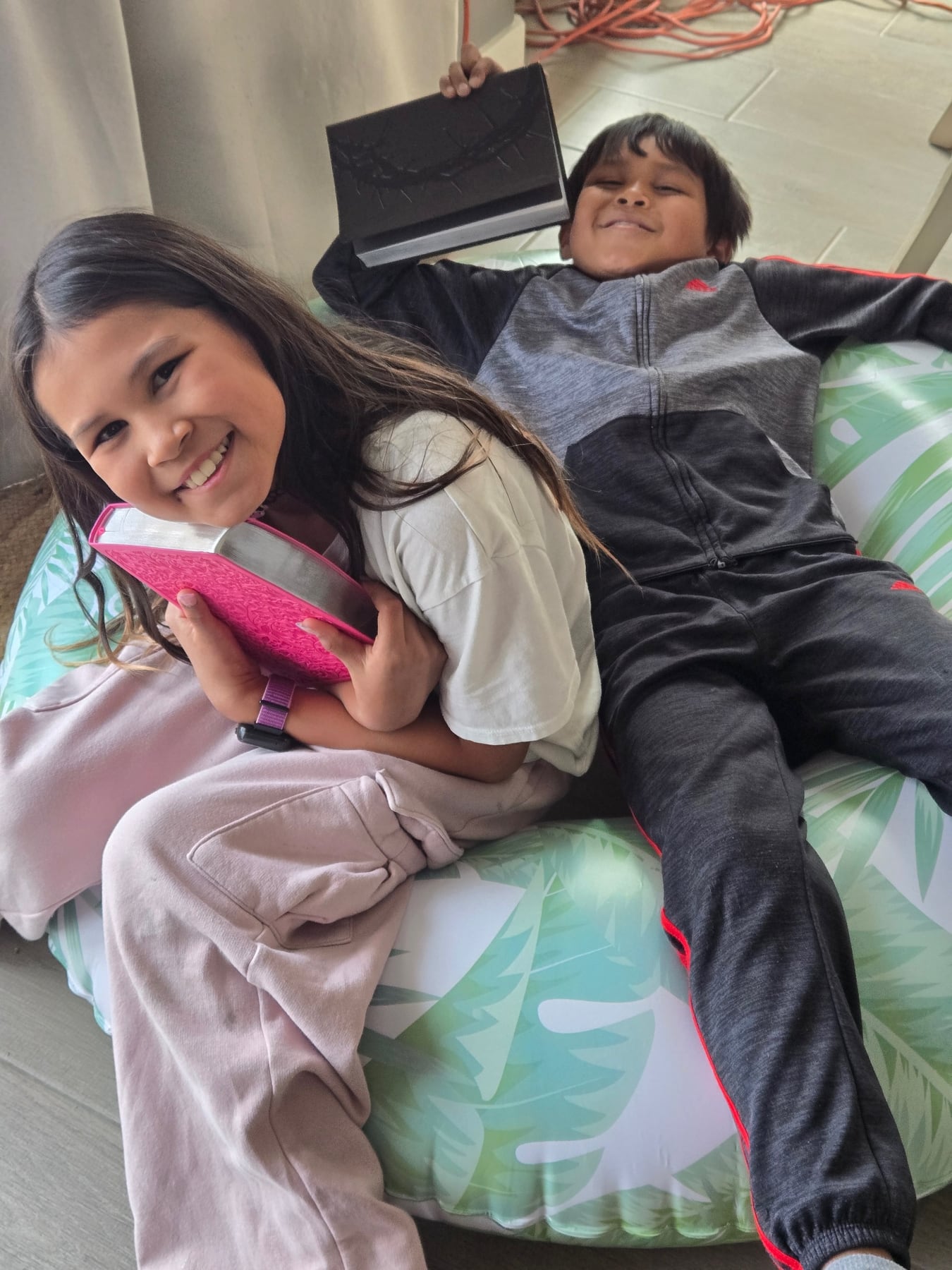 A girl and boy smiling together on a beanbag chair while holding books received through Foundation support